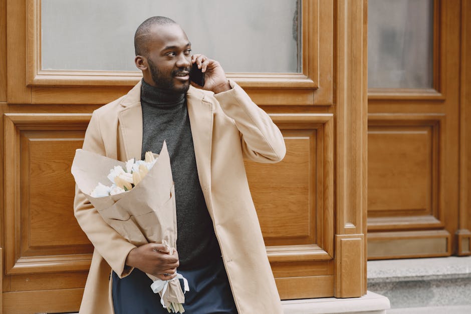 A man standing outside in front of wooden paneling, holding a bouquet of fresh white flowers wrapped in brown paper. He is dressed in a beige coat over a dark grey turtleneck, with short hair and a be